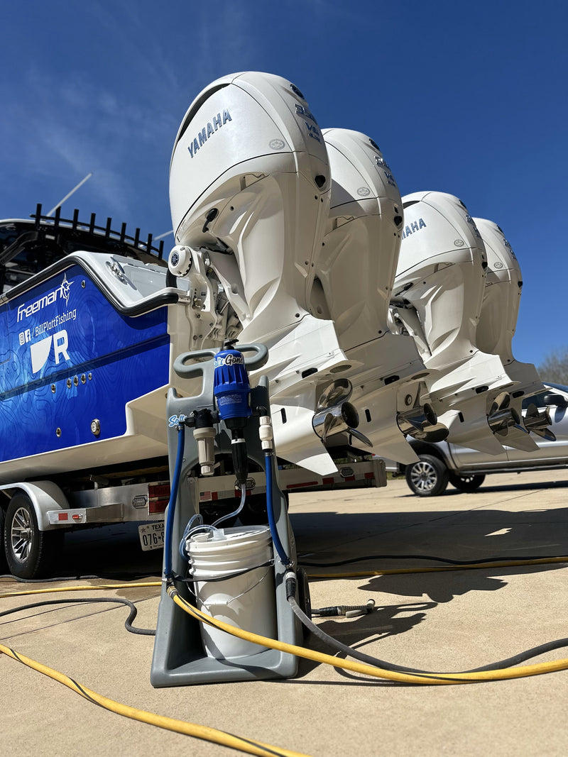 A close-up view of multiple Yamaha outboard motors on the back of a blue Freeman boat, with a Salts Gone® Cart with Injector Pump dispensing system set up in the foreground. The system includes a white 5-gallon bucket, hoses, and proportioning equipment. The scene is outdoors on a sunny day, with a clear blue sky in the background.